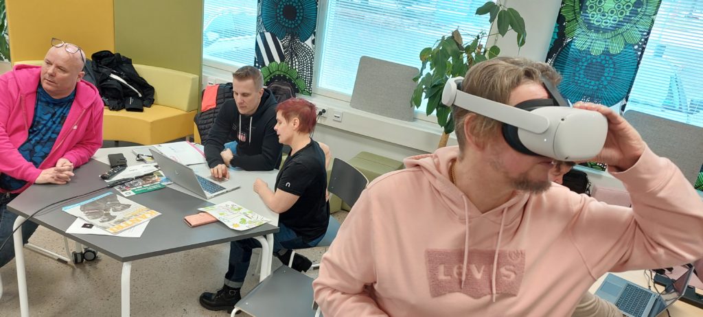 Yhdellä on VR-lasit. Kaksi henkilöä katsoo tietokonetta ja työskentelee sen edessä.