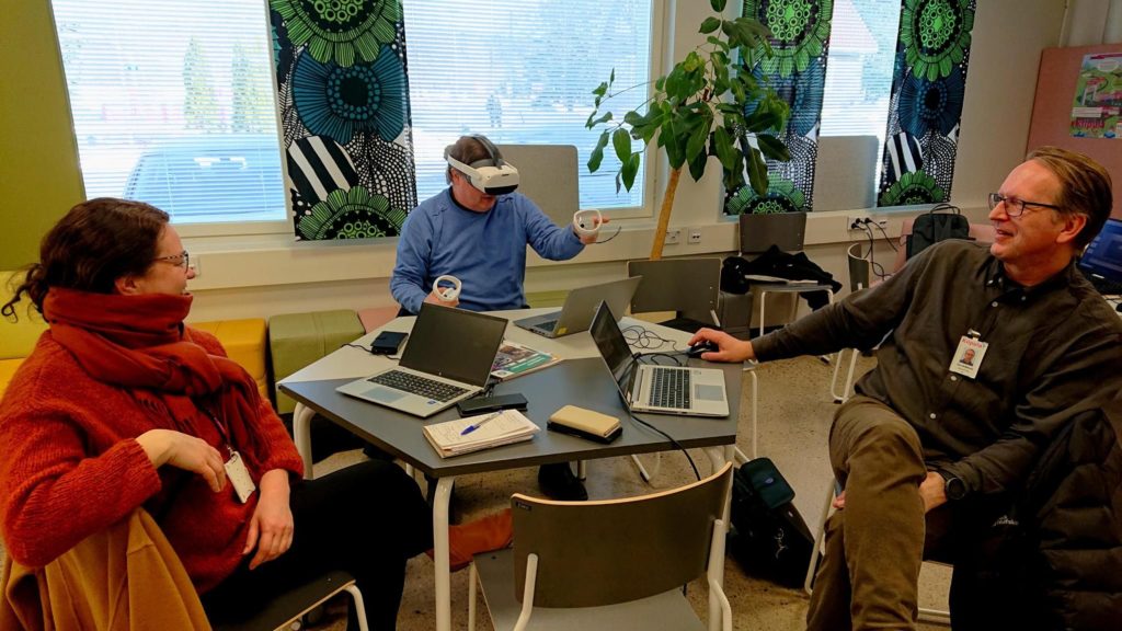 Kolme henkilöä istuu pöydän ympärillä ja yhdellä on VR-lasit päässä.