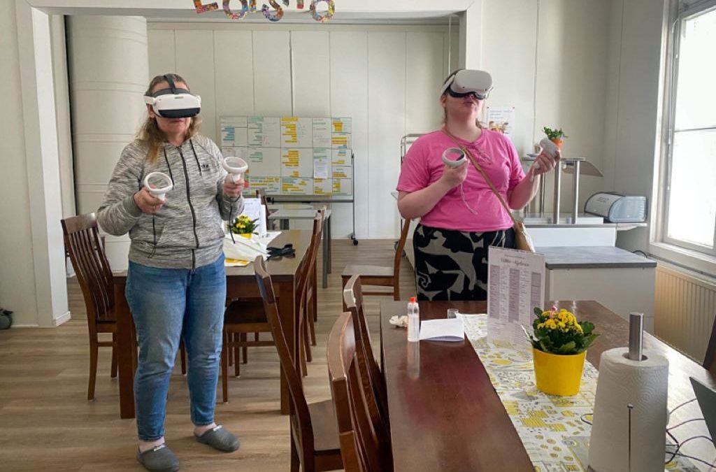 Kaksi henkilöä seisoo ruokailutilassa. Heillä on päässää vr-silmikot ja kädessään ohjaimet. Henkilöt pelaavat virtuaalista oppimispeliä.